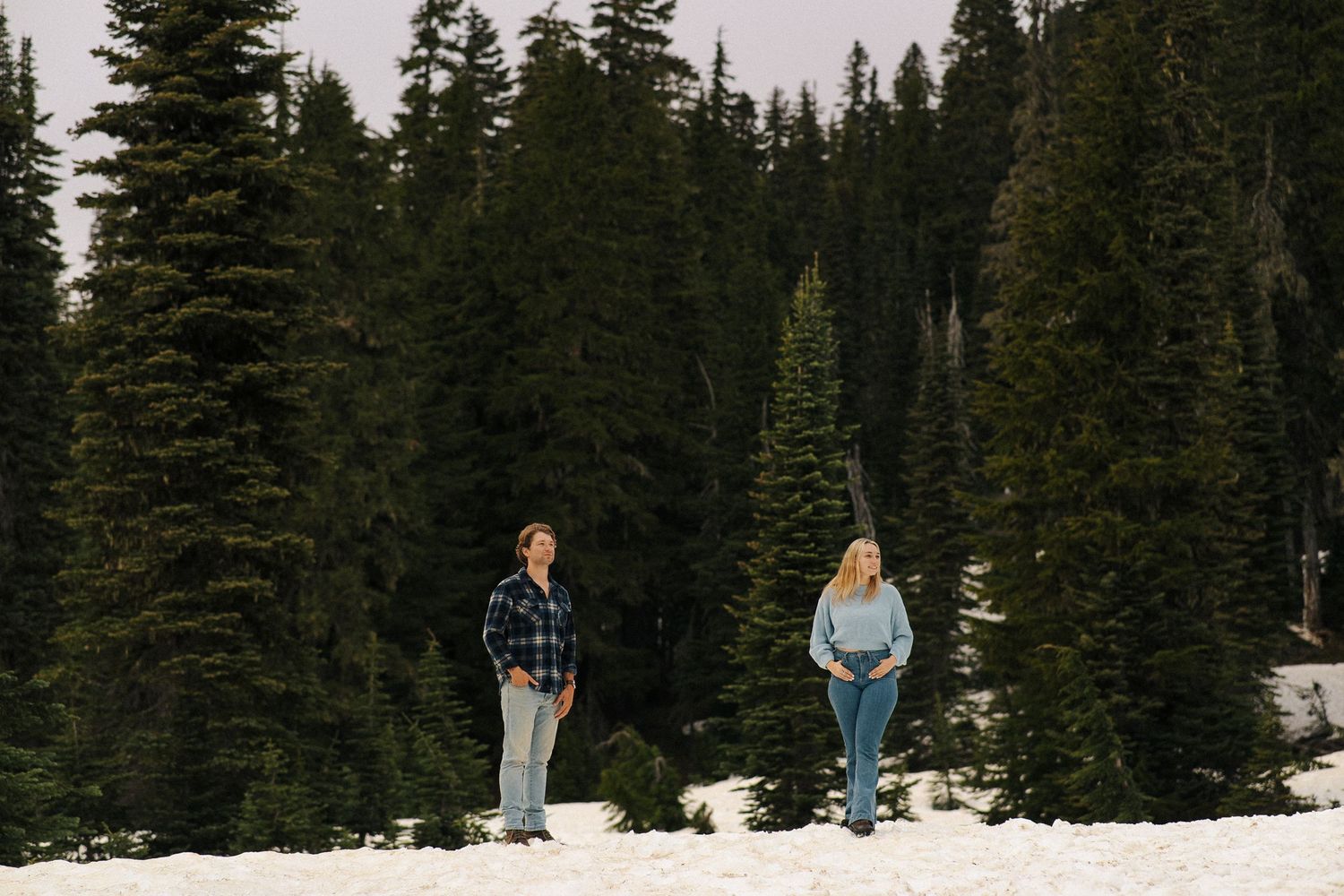 Two people stand in a snowy forest surrounded by tall evergreen trees in a winter mountain landscape.
