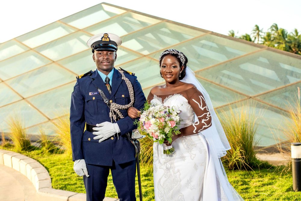 Wedding couple poses outdoors in elegant attire with white dress and military uniform holding a bridal bouquet.