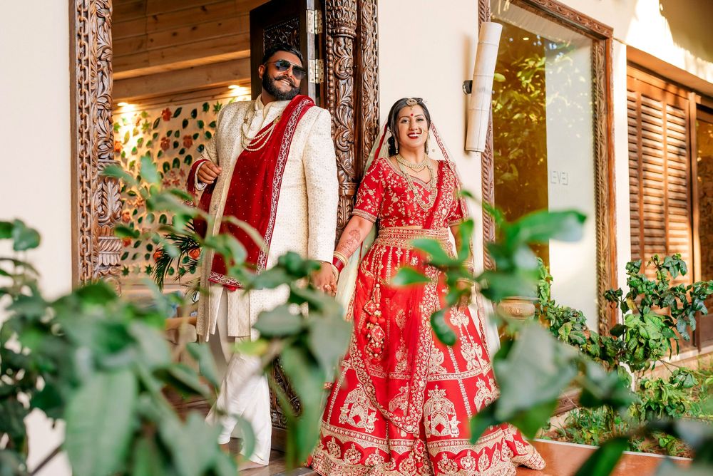A couple in traditional Indian wedding attire pose together on a wooden porch surrounded by greenery.