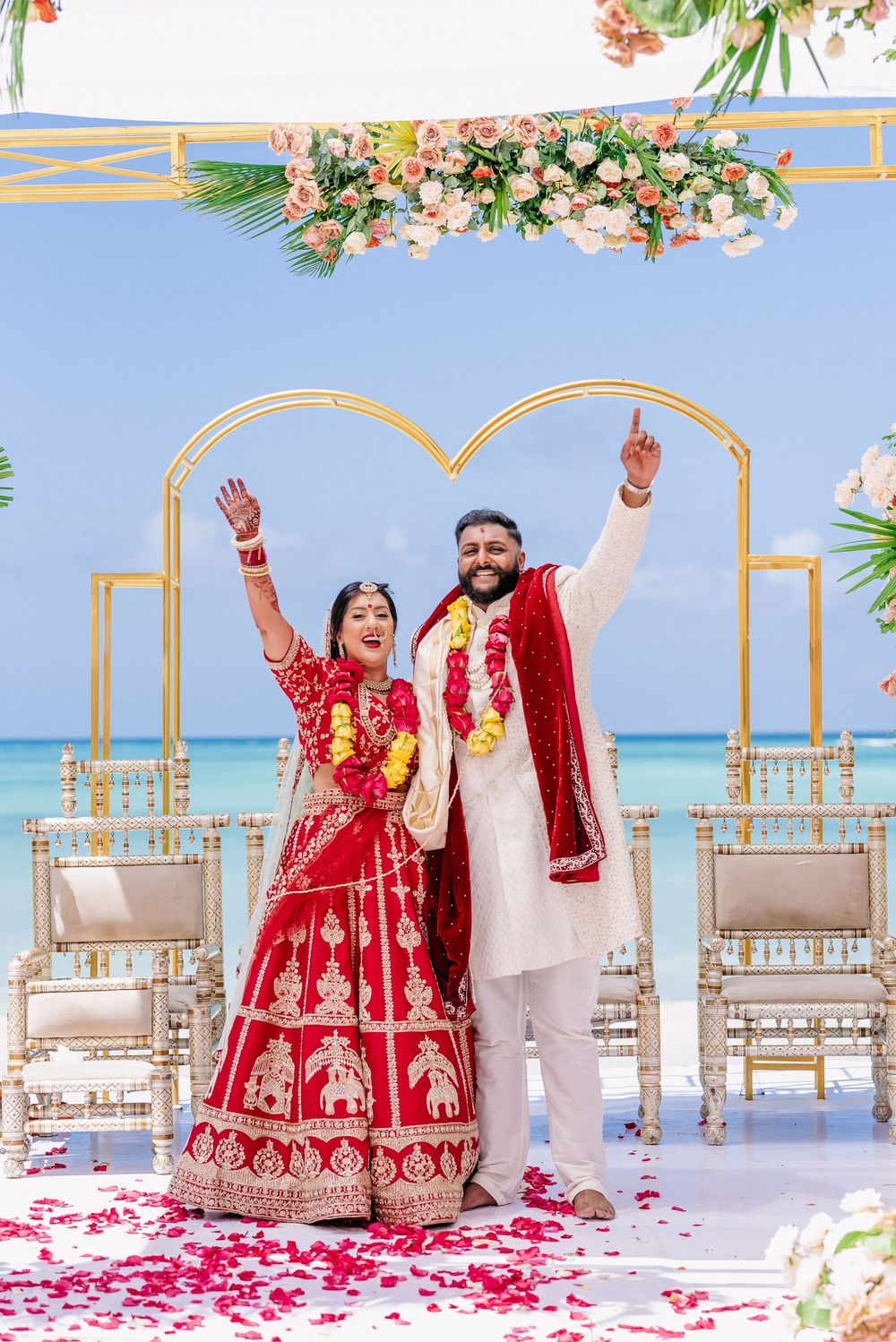 Traditional Indian wedding ceremony under floral arch with ocean backdrop.