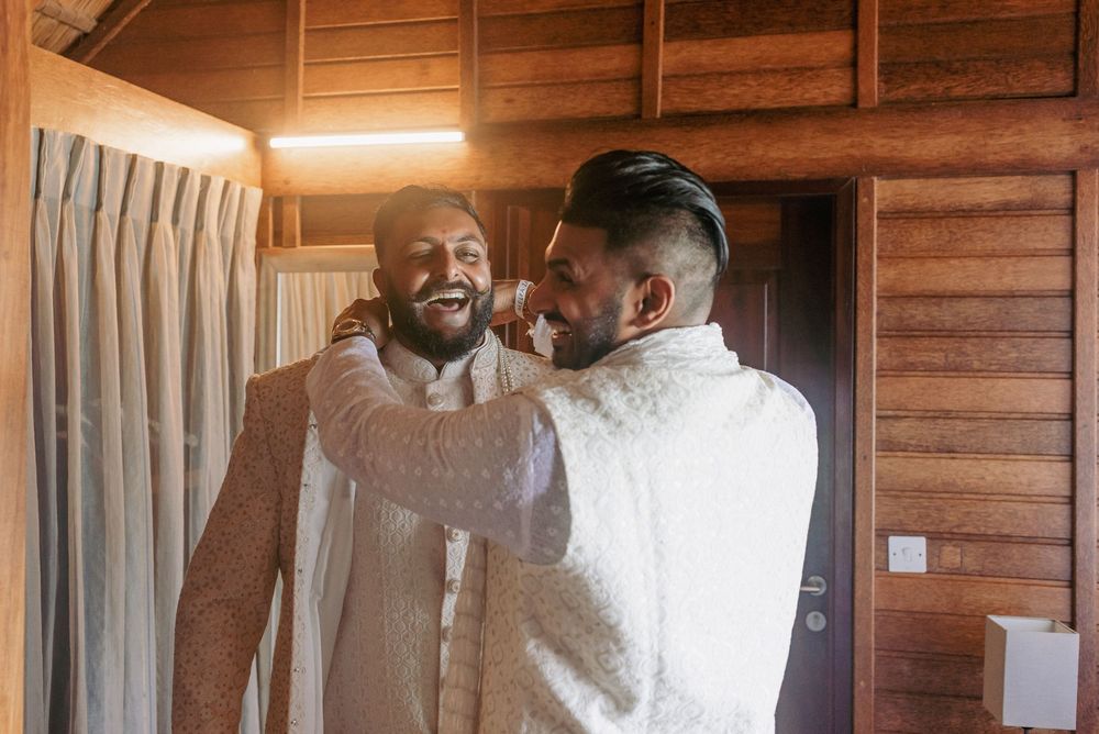 Groomsmen in white traditional attire share laughs and hugs in a wooden cabin setting.