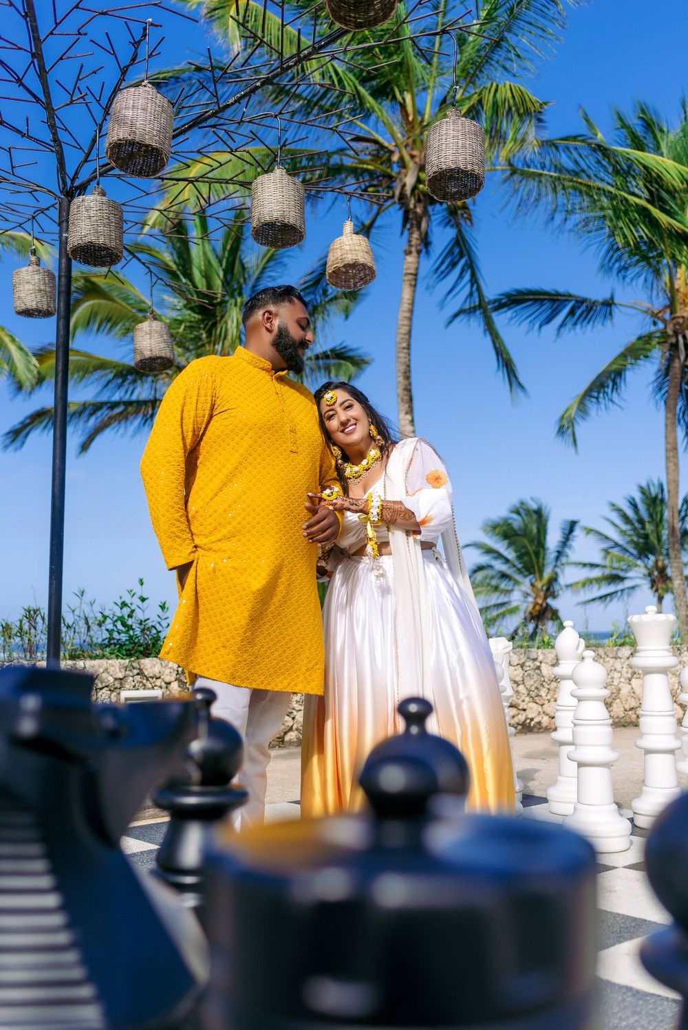 Romantic couple in traditional attire poses by giant chess set with palm trees.