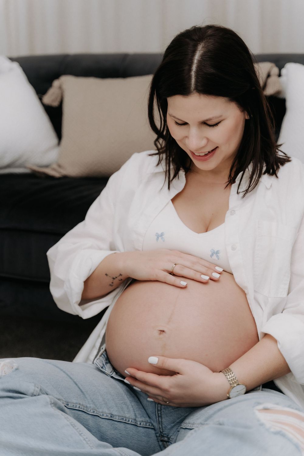 Schwangere Frau sitzt auf dem Sofa und hält mit beiden Händen liebevoll ihren nackten Babybauch