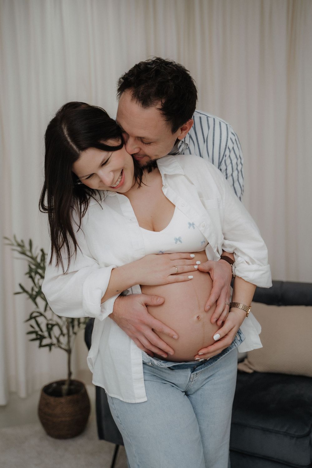 Paar umarmt sich innig bei einem Babybauchshooting im Wohnzimmer, beide lächeln und halten den Bauch