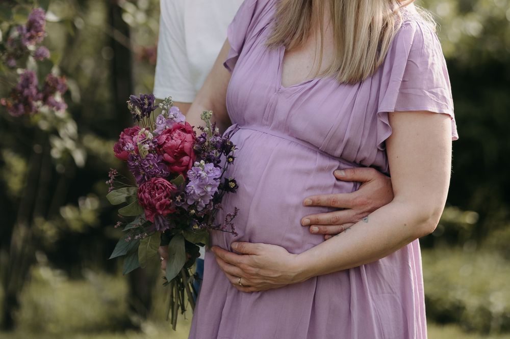 Paar im Garten hält gemeinsam den Babybauch, die Frau trägt ein fliederfarbenes Kleid und hält einen Blumenstrauß