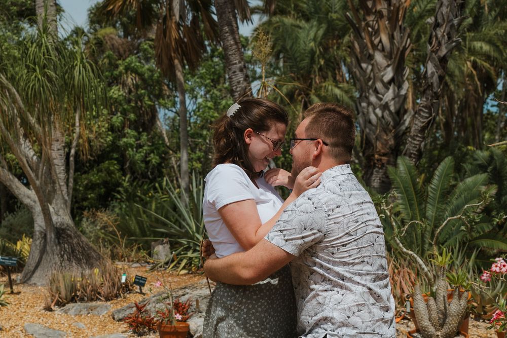 A romantic couple shares an intimate moment in a tropical garden setting surrounded by palm trees and lush vegetation.