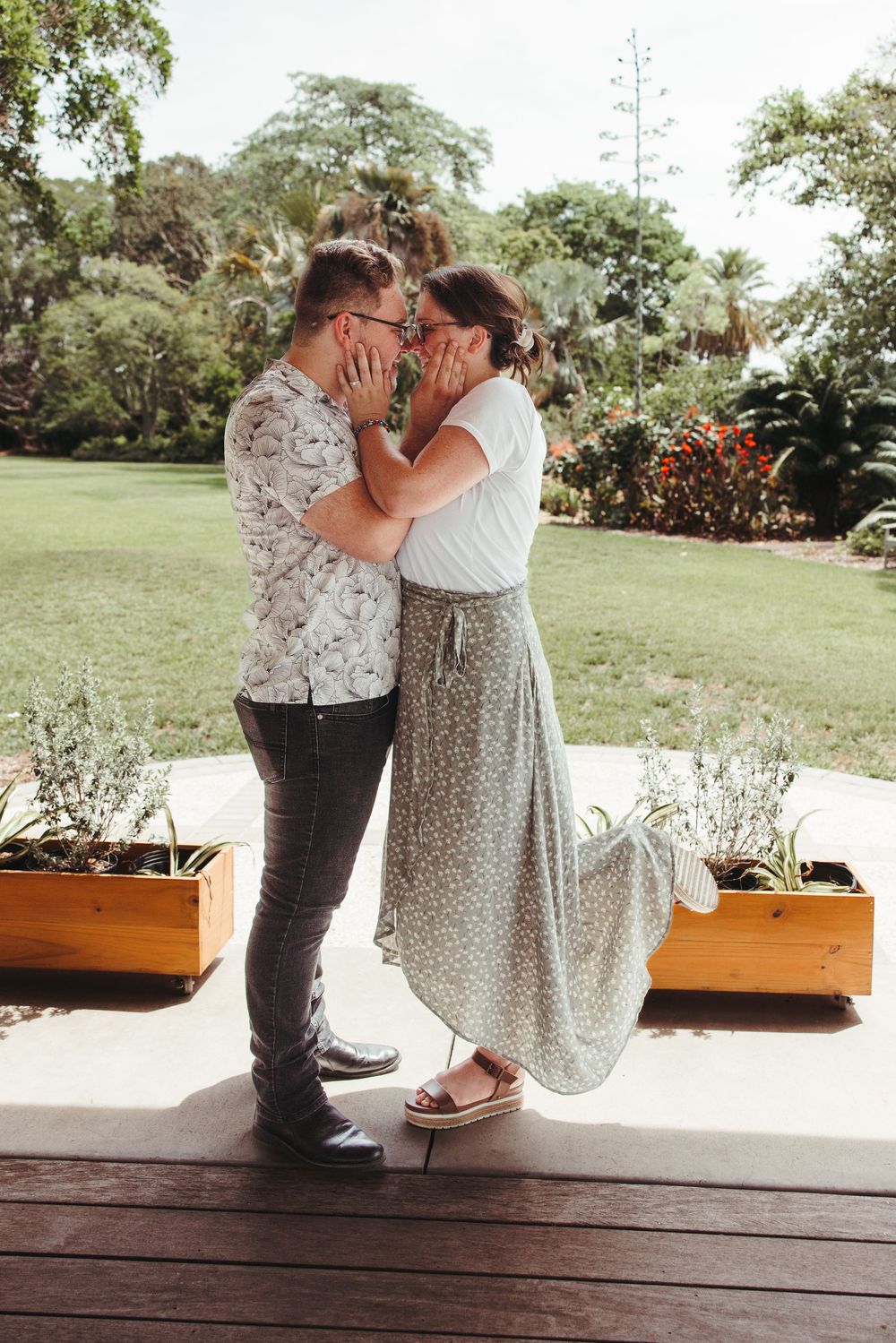 Two people in romantic poses share sweet moments on a wooden deck surrounded by lush greenery.