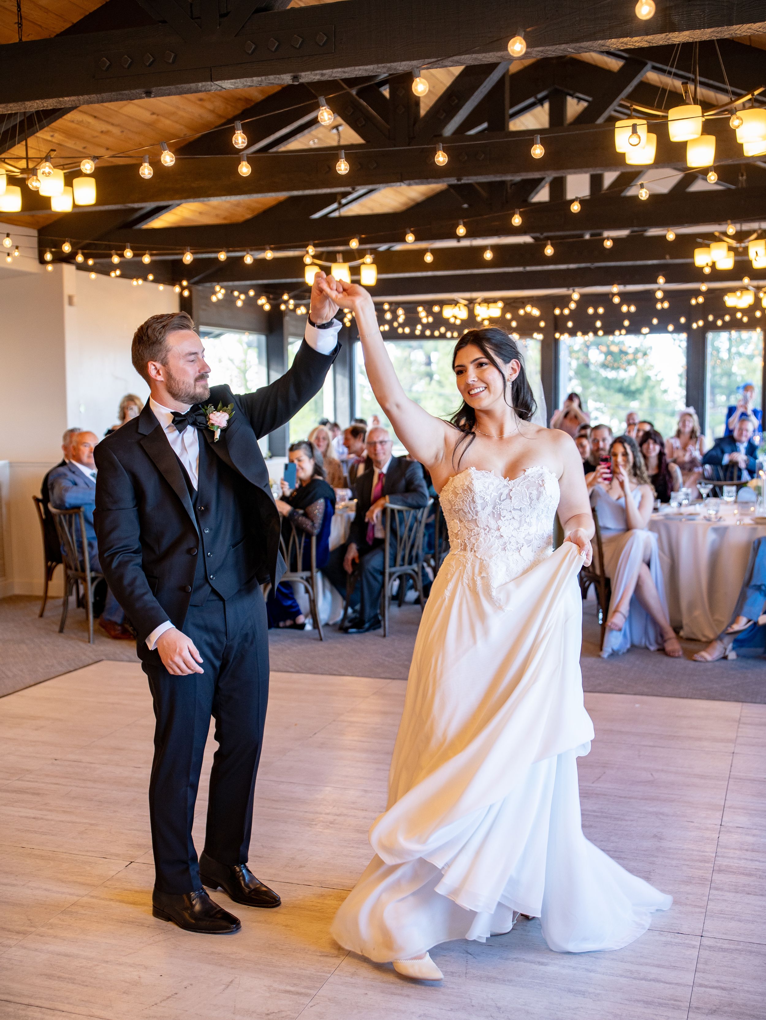 A joyous wedding celebration with dancing newlyweds under string lights in an elegant rustic-style indoor reception venue.