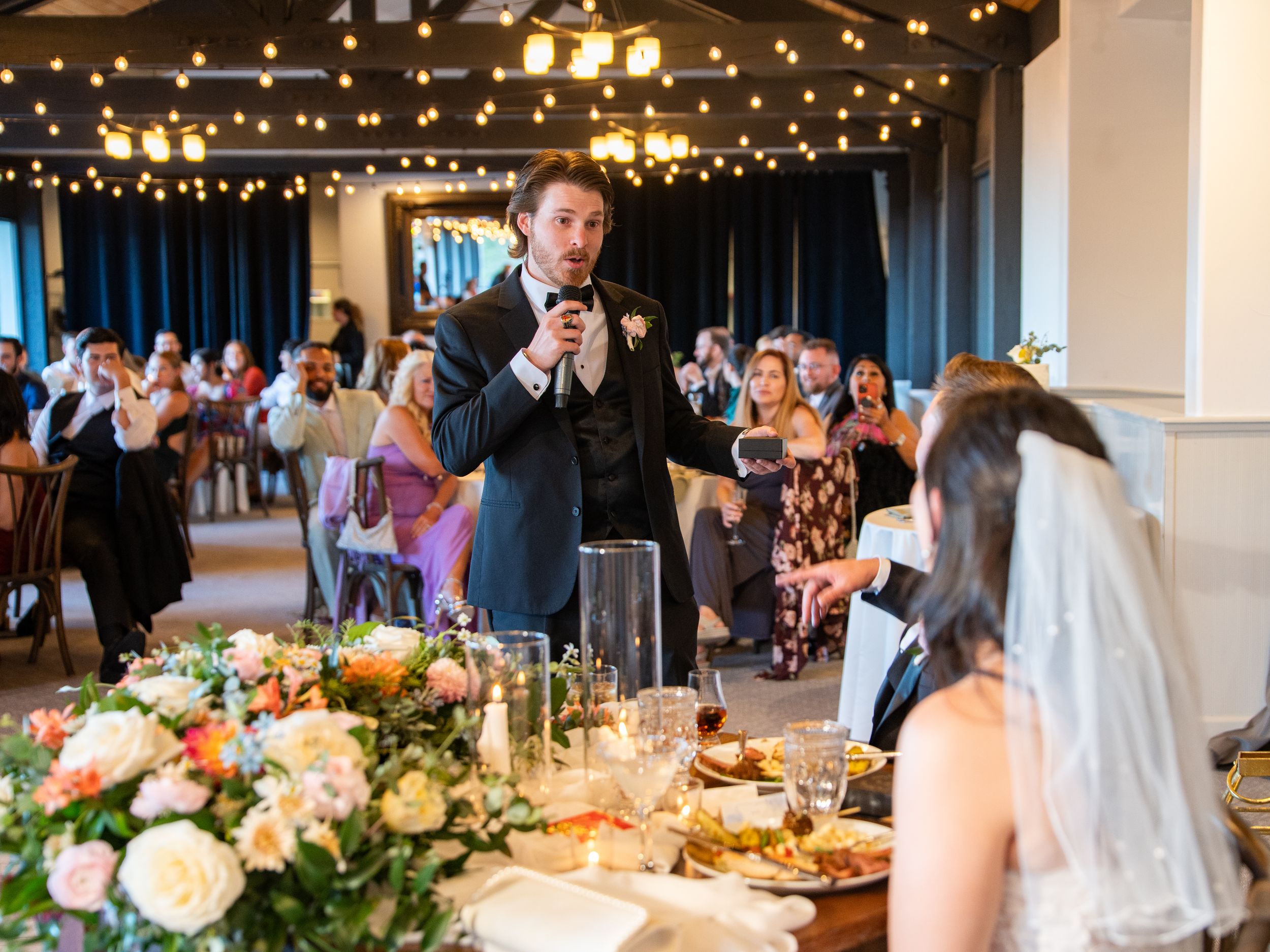 A wedding reception hall decorated with string lights and elegant floral arrangements features guests dining at round tables.