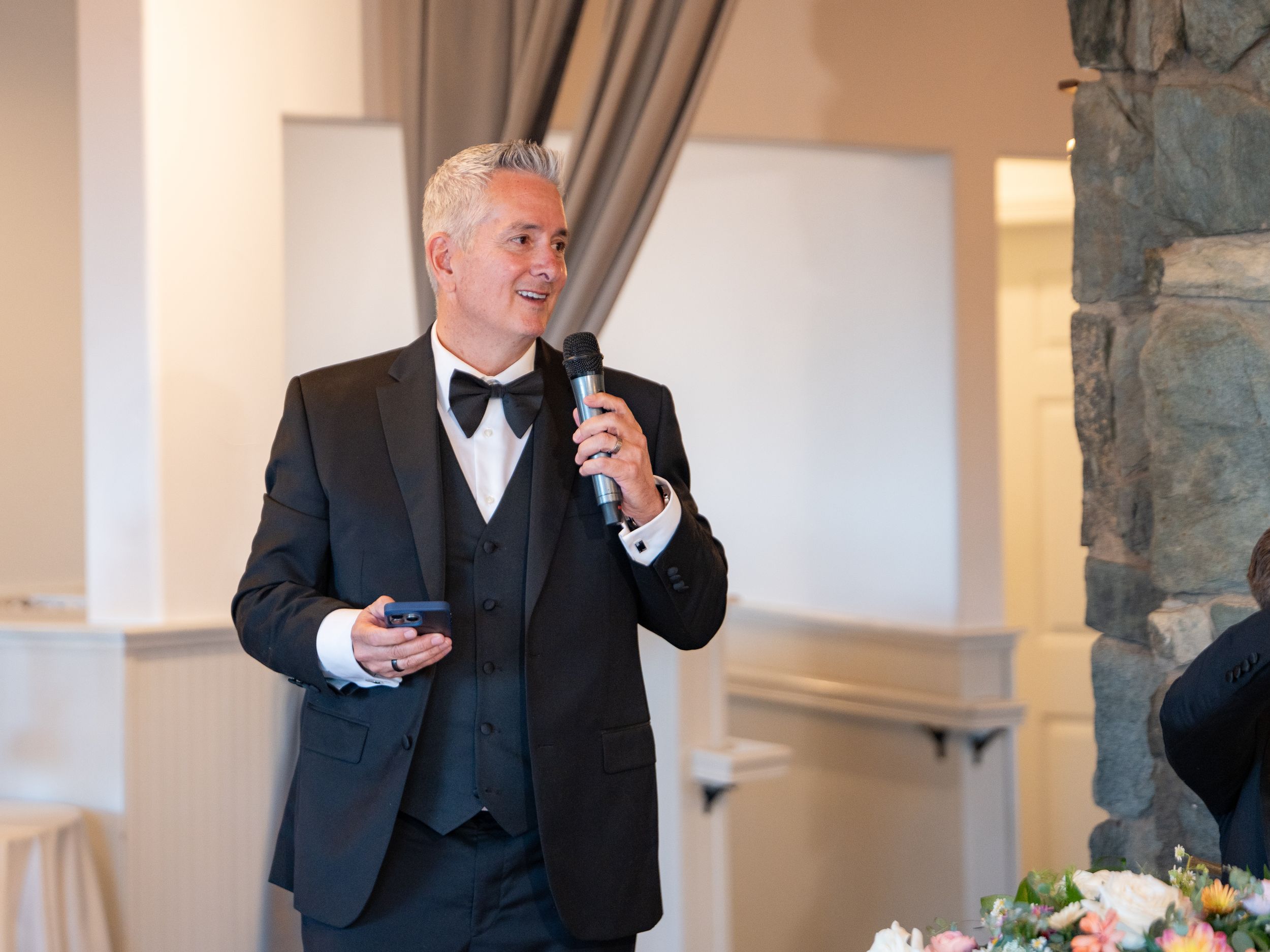 Well-dressed man in a tuxedo gives a toast or speech with a microphone at an elegant event venue.