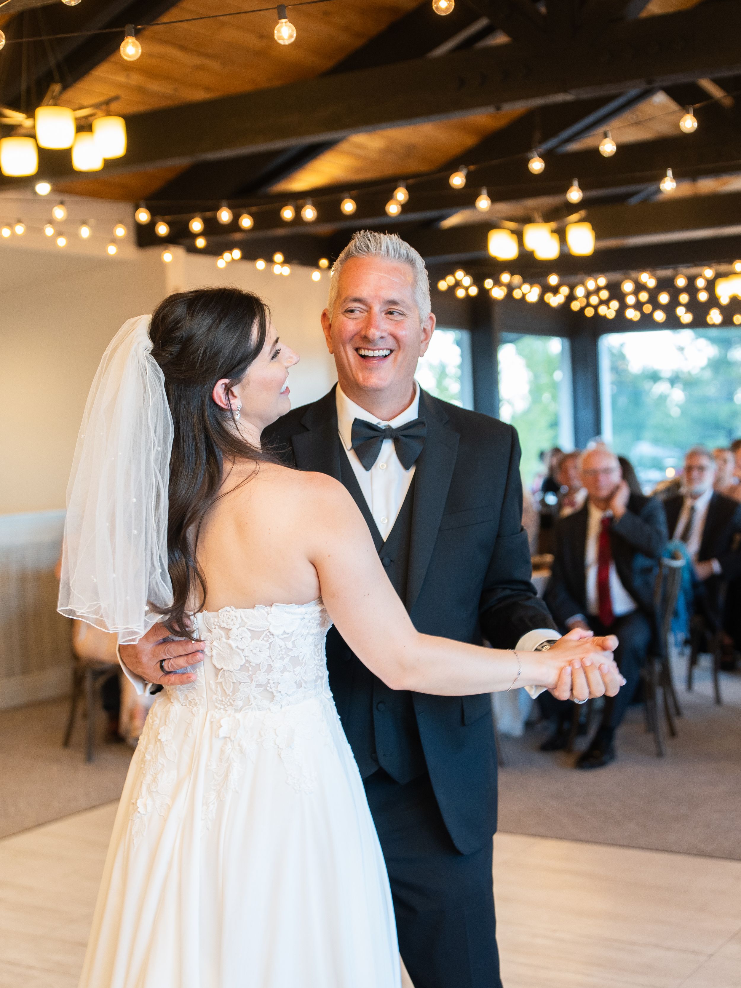 A bride in a white strapless gown shares a dance with a companion in a black tuxedo under romantic string lighting.