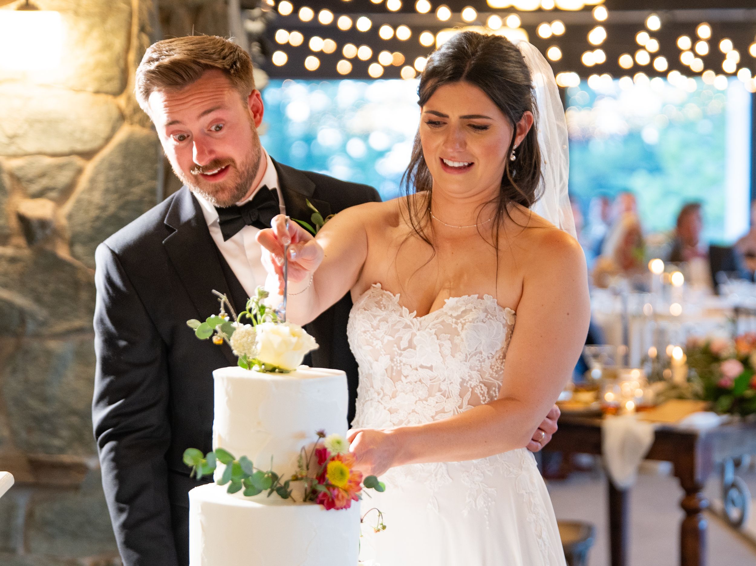 A newlywed couple cuts their elegant white wedding cake together under decorative string lights at their reception.