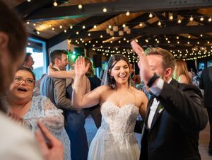 A wedding reception party celebrates on a dance floor under warm string lights and decorative ceiling beams at an indoor venue.