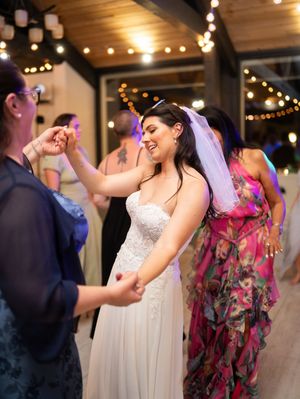 A lively dance celebration at an indoor wedding reception with decorative string lights and wooden beams overhead.