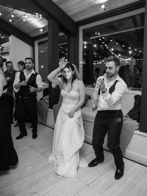 Wedding guests dance joyfully together at an indoor reception with wooden beams and string lights visible in background.