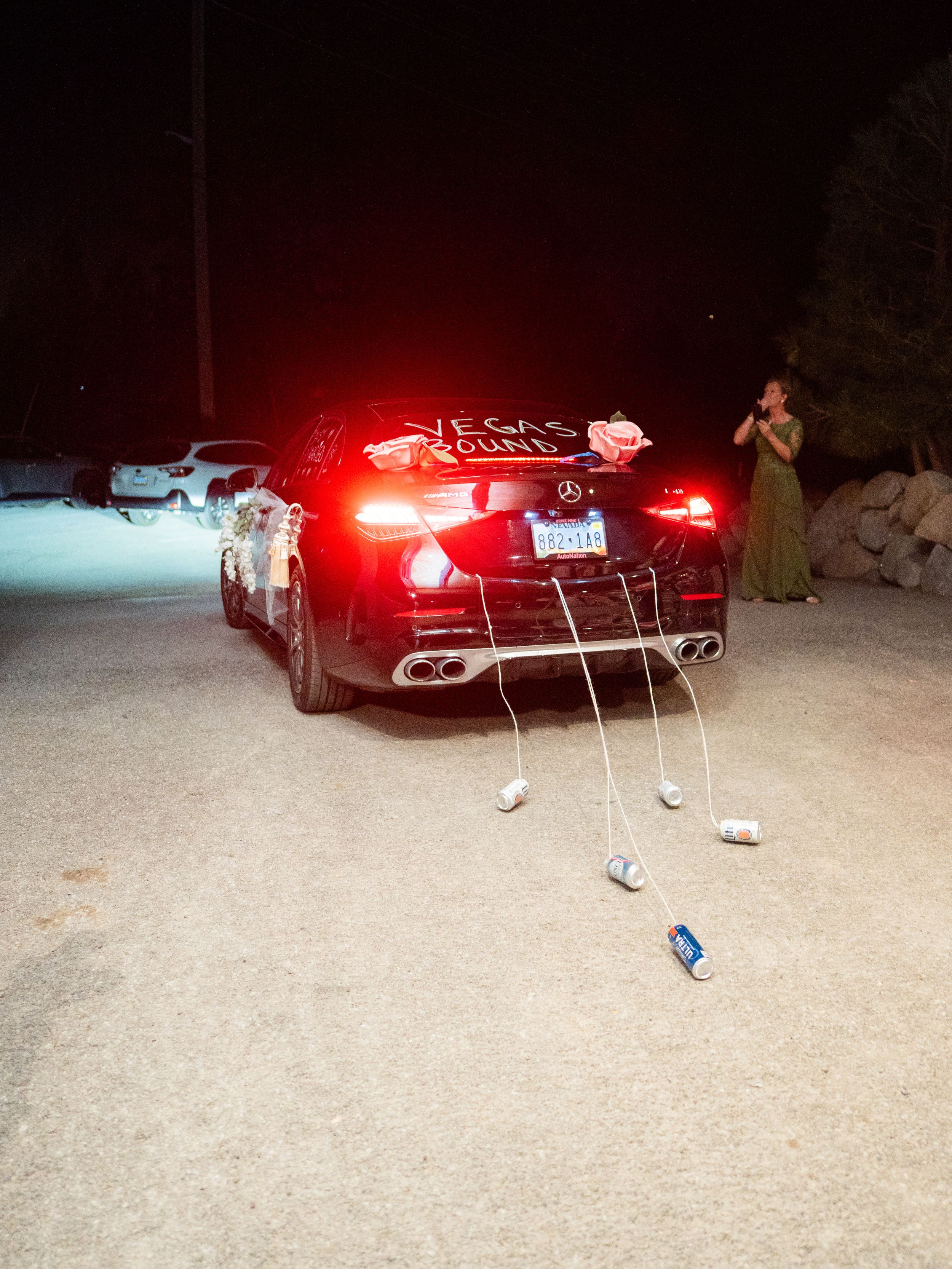 A black car with cans tied to the back bumper at night with brake lights glowing red in the darkness.