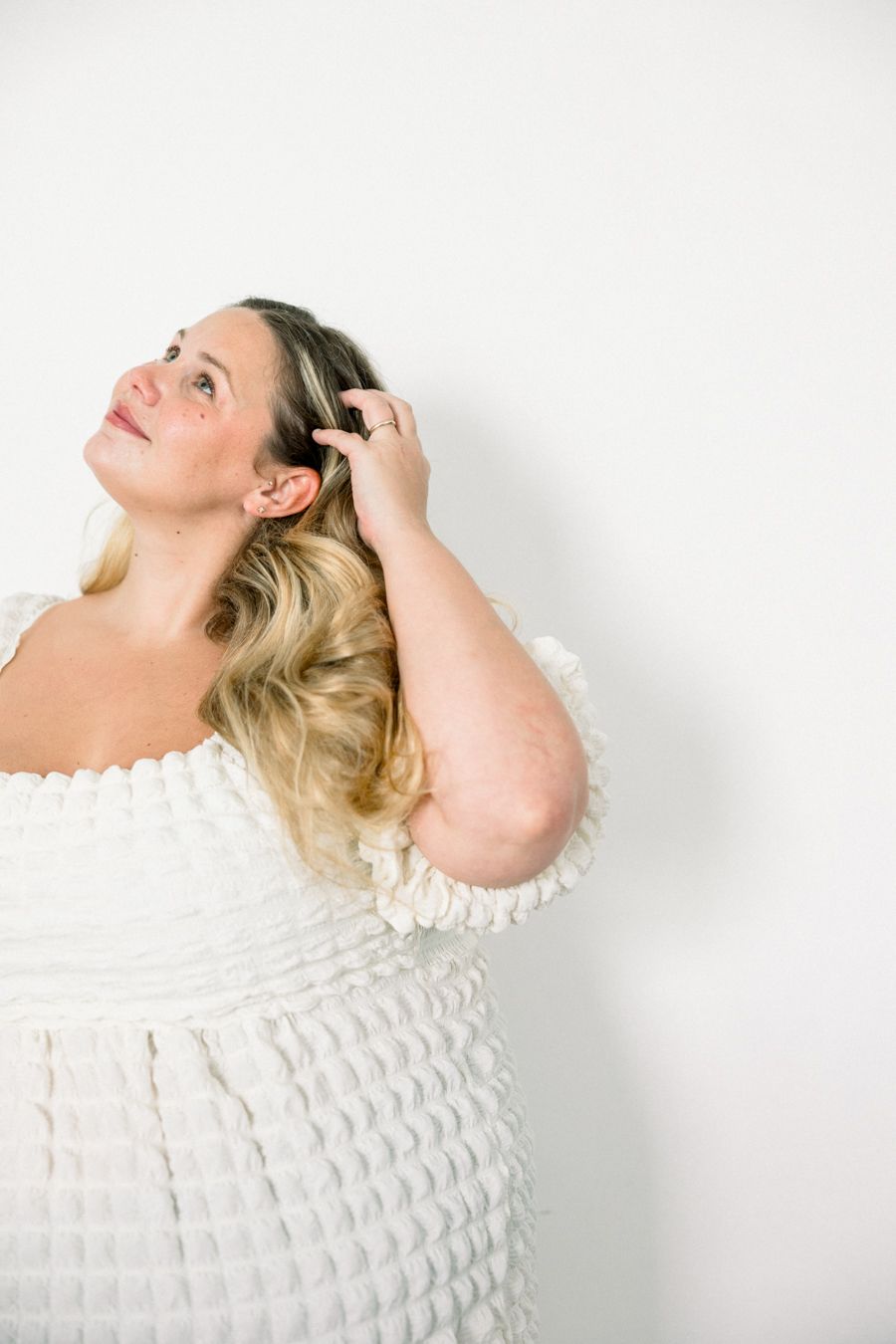 Woman in white textured off-shoulder dress poses against plain wall with hand in blonde wavy hair. Best Augusta Newborn Photographer
