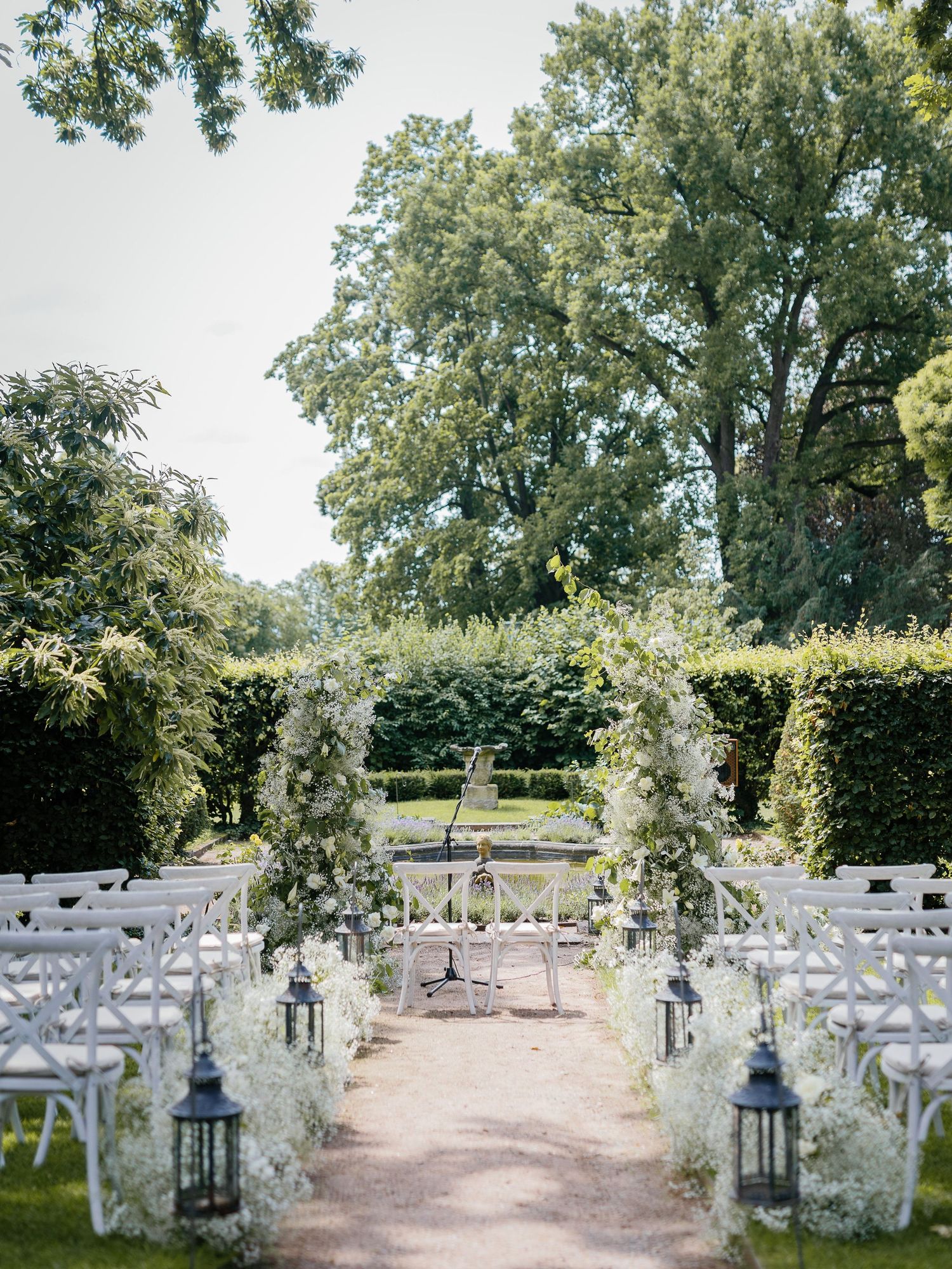 Elegant outdoor wedding ceremony setup with white chairs, floral arches and black lanterns lining a garden path.