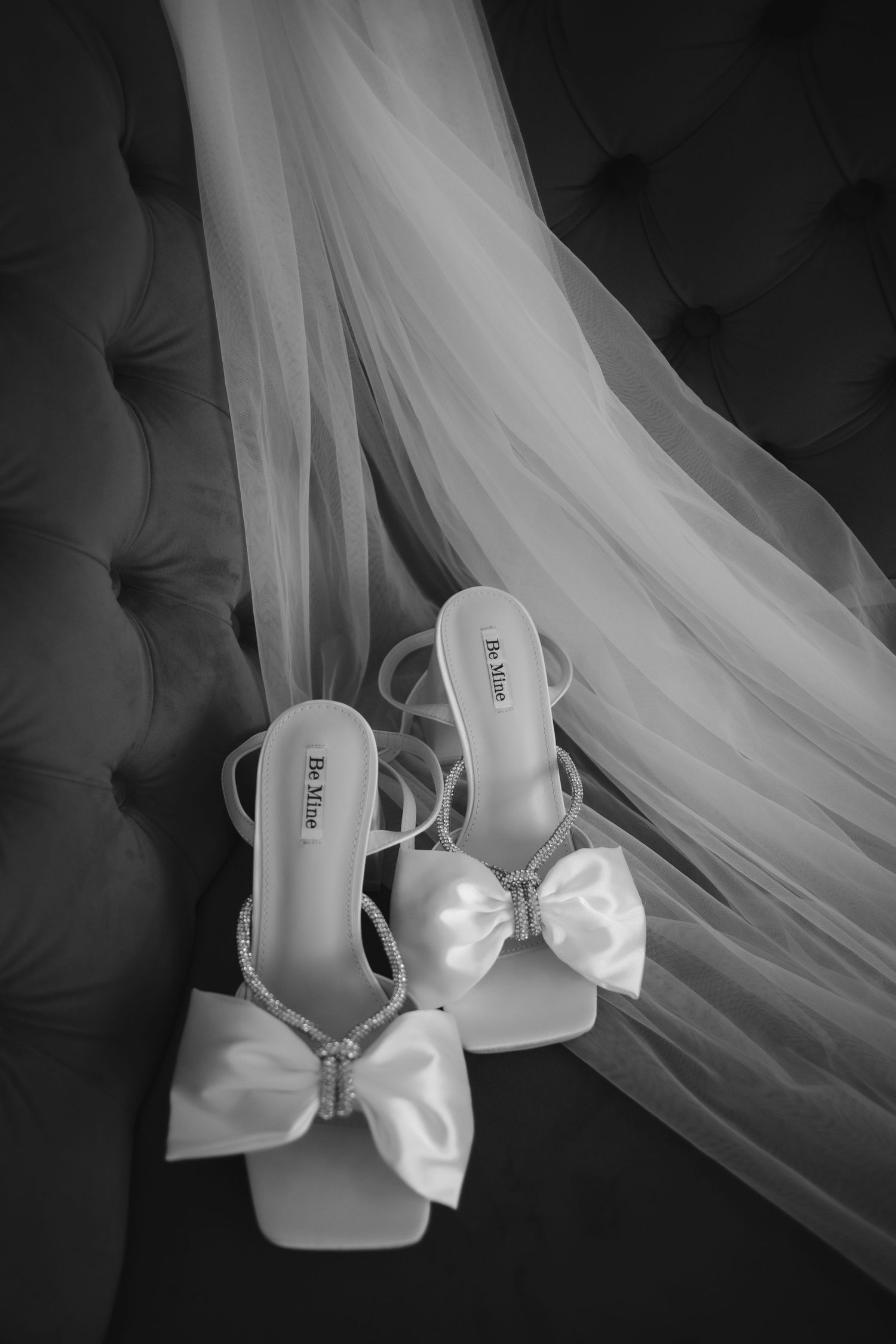 Black and white wedding photo of elegant bridal shoes with satin bows and veil on tufted fabric background.
