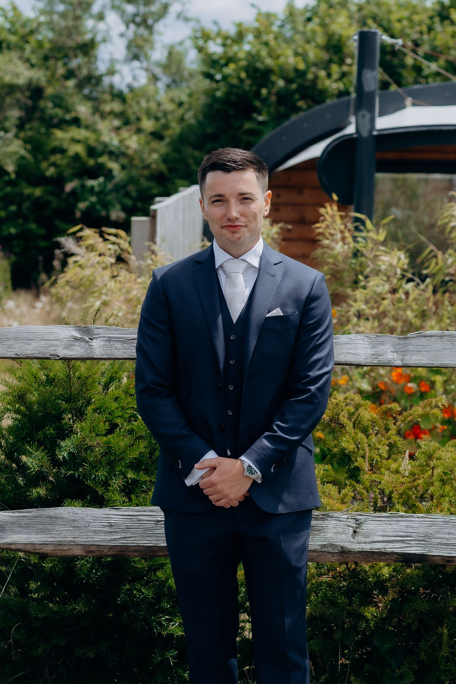 Someone wearing a navy blue three-piece suit and white tie poses outdoors against a rustic wooden fence and garden backdrop.