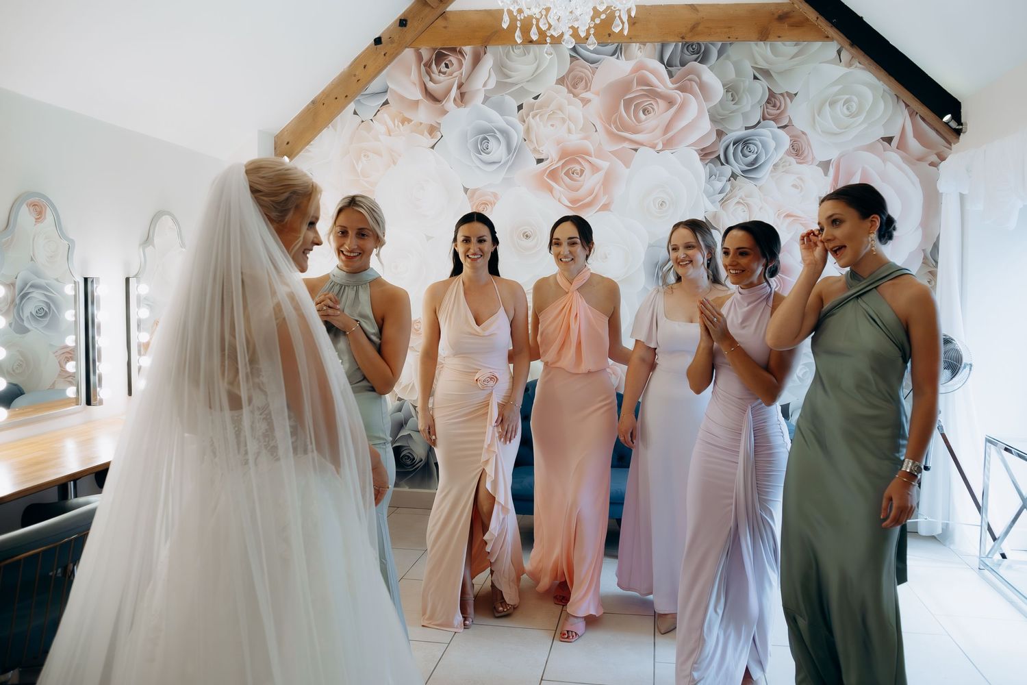 A bridal party shares a joyful moment in a dressing room decorated with pink and silver floral backdrop.