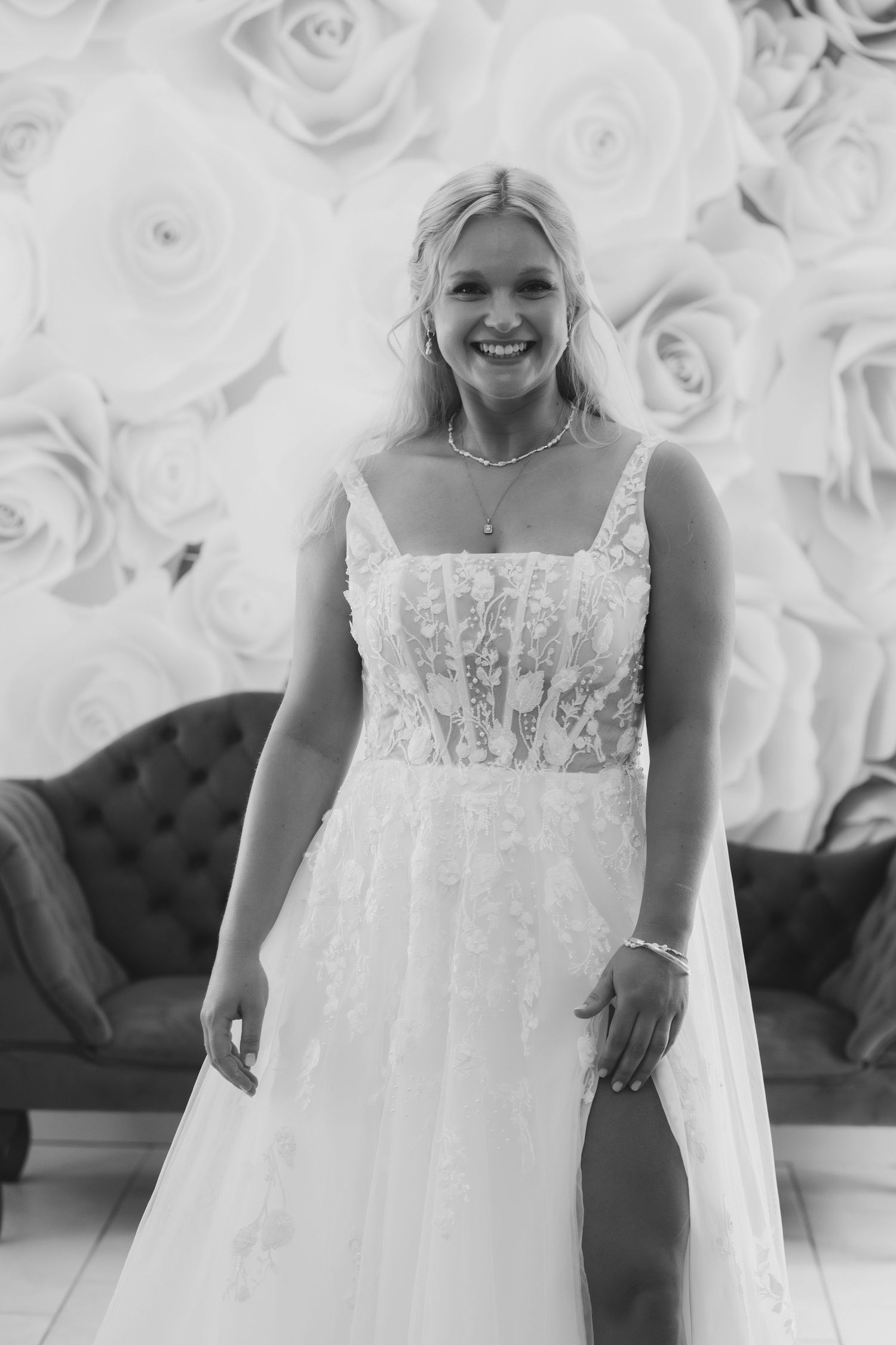 Black and white photo of someone wearing an elegant lace and tulle wedding gown against a floral backdrop.