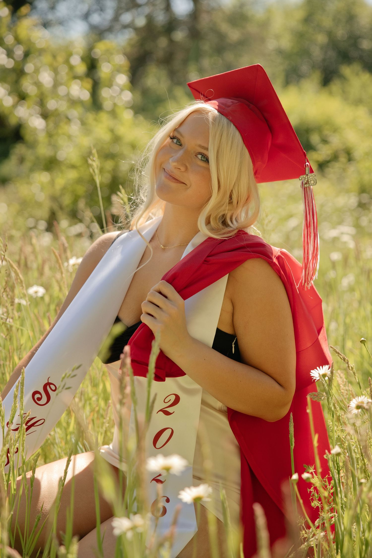 A graduate in a bright red cap and gown leans on a concrete pillar outdoors with curly hair and a cheerful expression.