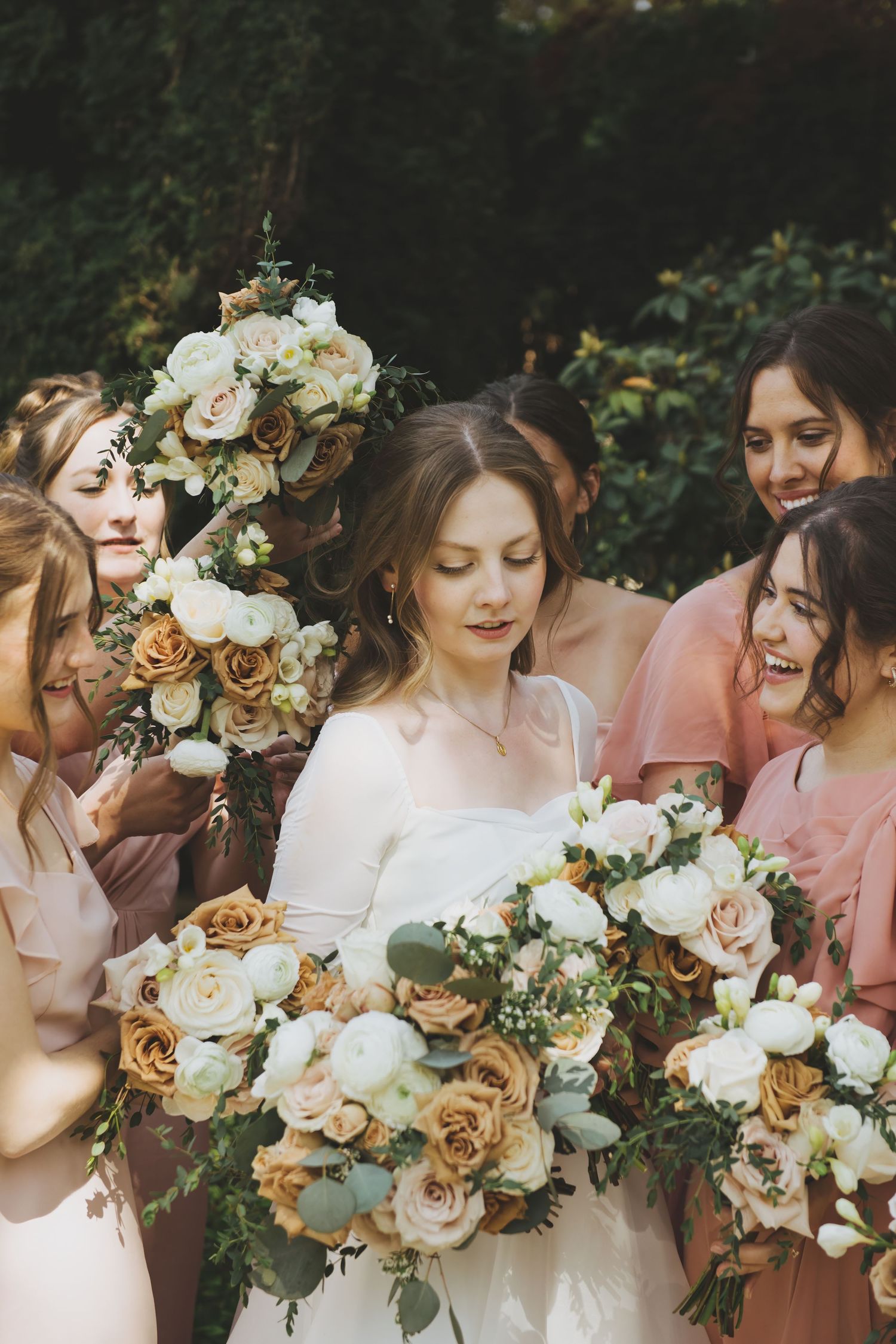 Bride and bridesmaids in soft pink dresses share a joyful moment while holding romantic bouquets of white and caramel roses.