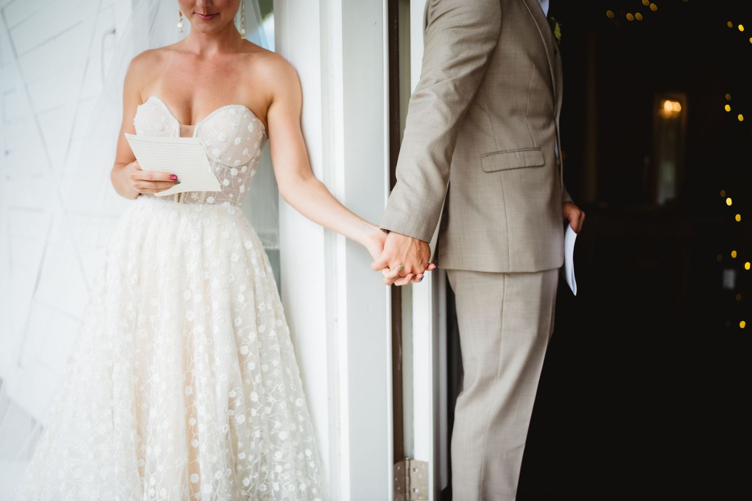 A bride in a white lace wedding dress holds hands with her groom wearing a light gray suit at a doorway.