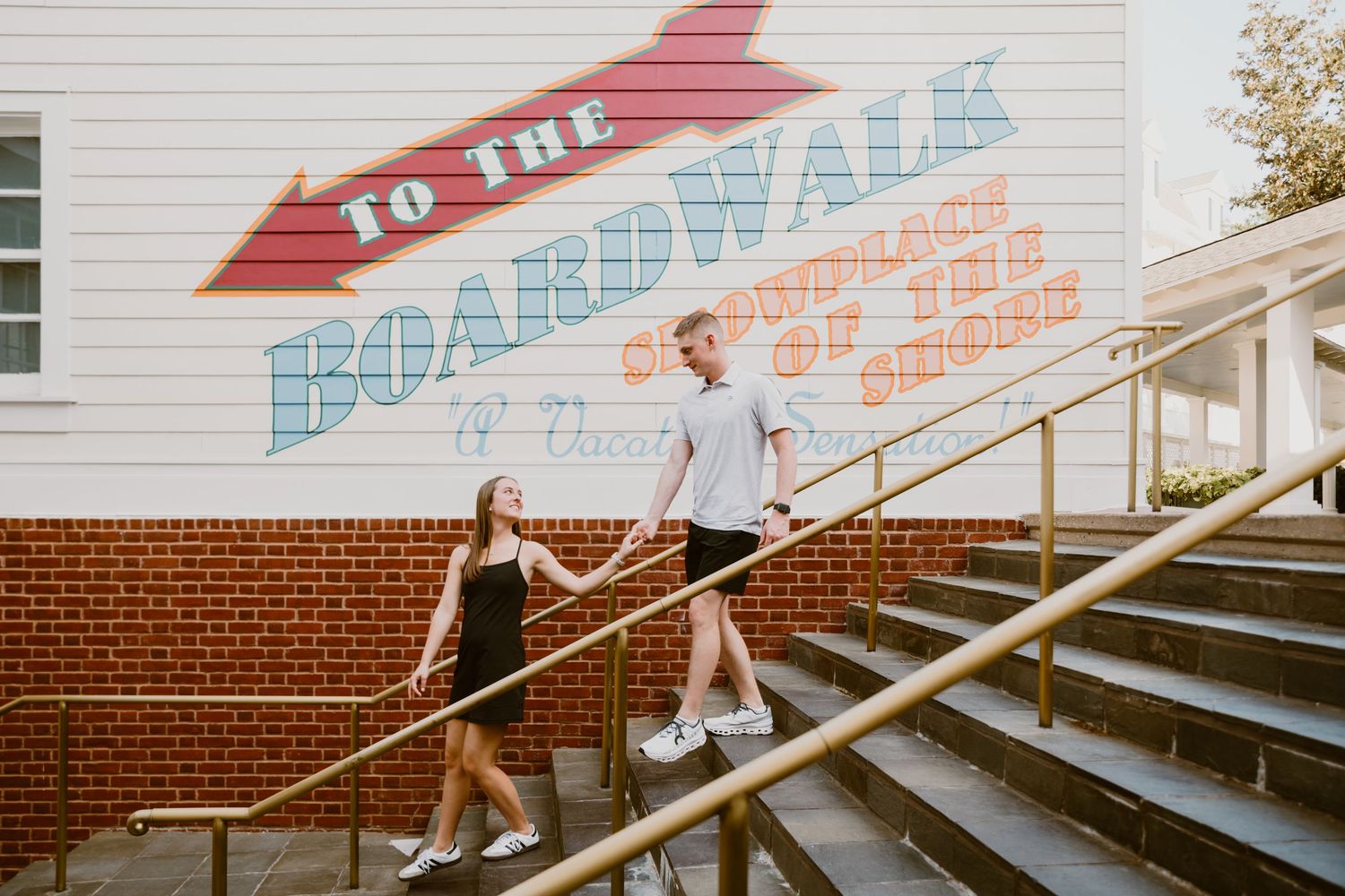 Young couple running at "To The Boardwalk" mural stairs at Disney Boardwalk during surprise proposal. Cred: LexTakePix