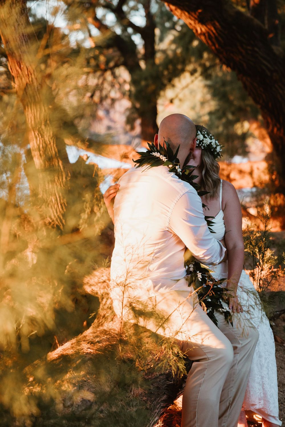 Couple in wedding attire share an intimate moment bathed in golden sunset light through trees with greenery decorations.