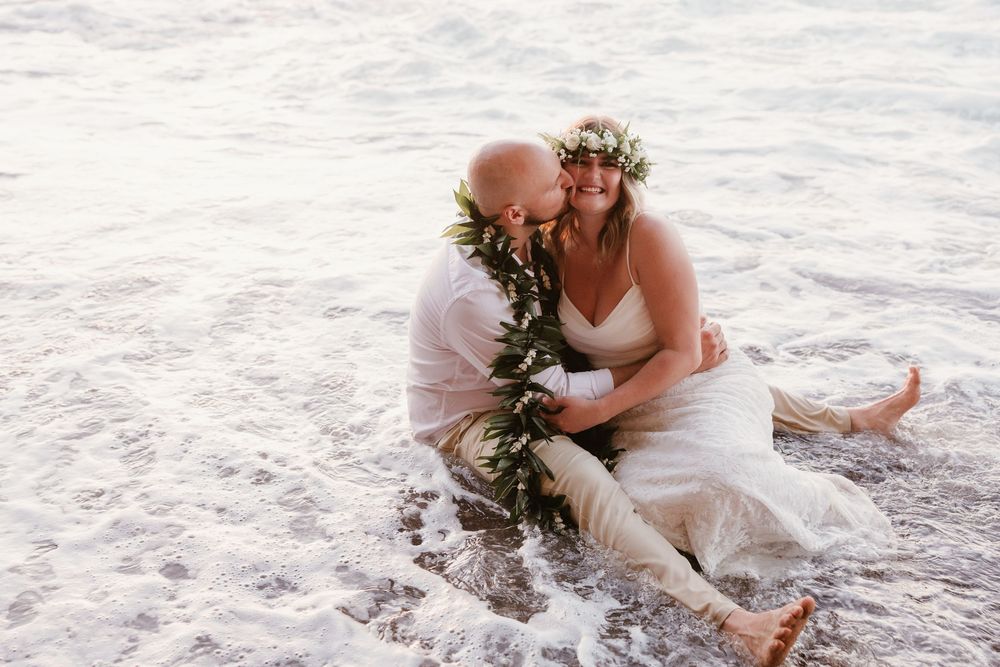Two people sit together on sandy beach shore with waves and sunlight in background.