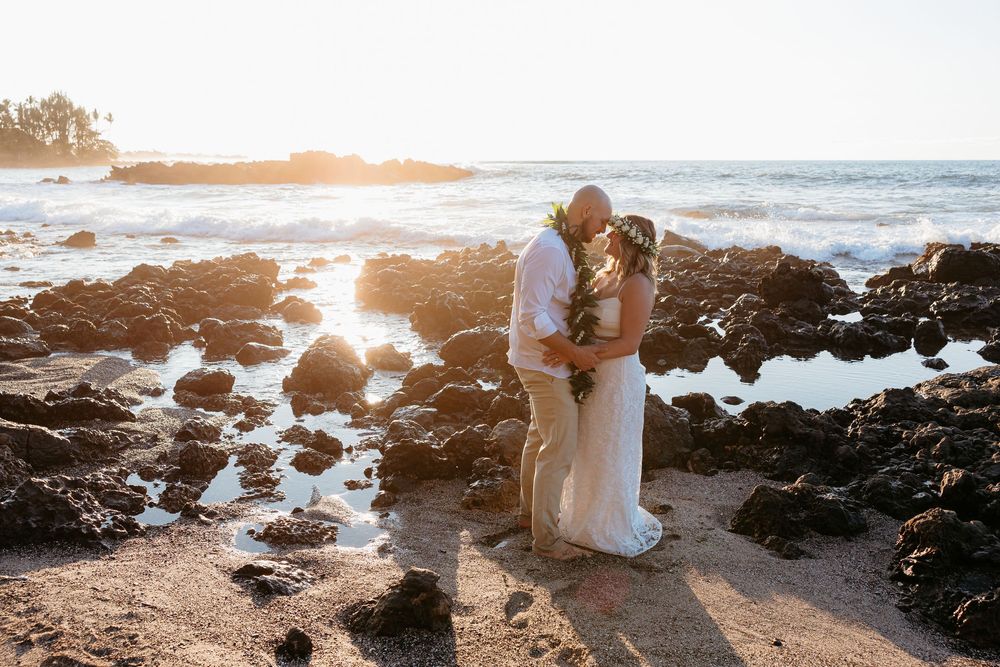 Two figures hold hands at sunset on a rocky coastline as ocean waves crash against volcanic rocks in Hawaii.