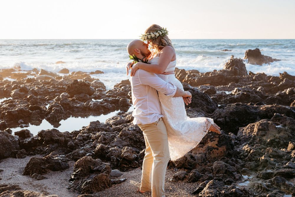 Romantic couple embraces on rocky beach at sunset with ocean waves crashing in background.