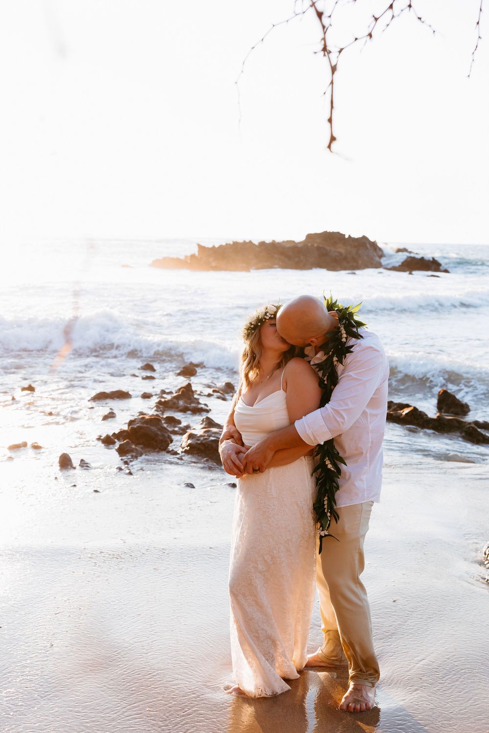 A couple embraces romantically on a sandy beach with ocean waves and rocks in the background at sunset.