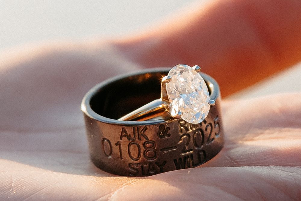 Black wedding band with engraved text and large diamond solitaire ring on pink fabric in soft lighting.