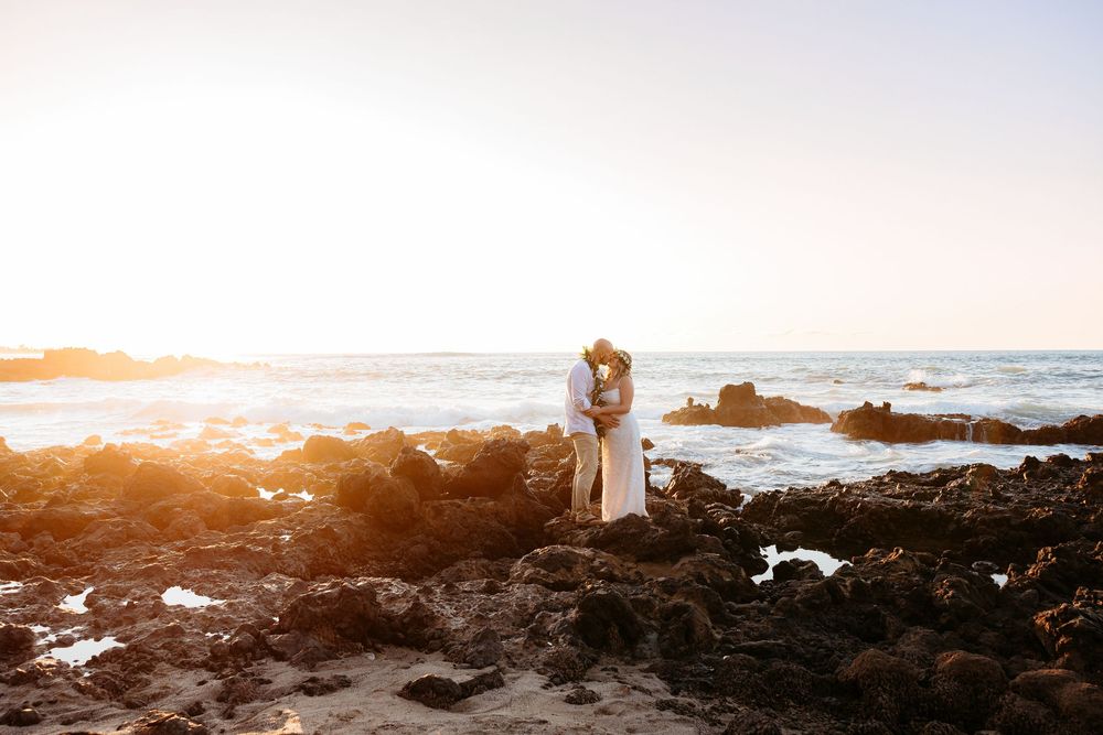Couple embraces on rocky beach at sunset while ocean waves crash against volcanic rocks in Hawaii.
