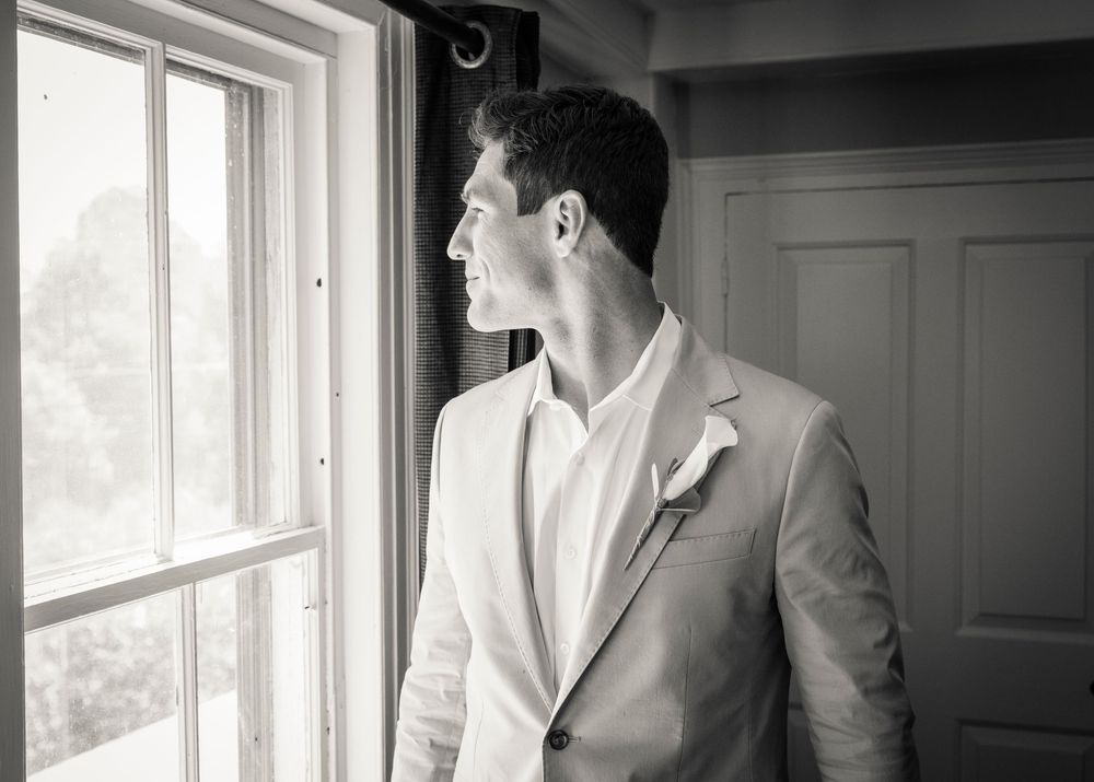 Black and white photo of a person in a light suit jacket gazing thoughtfully out a bright window.