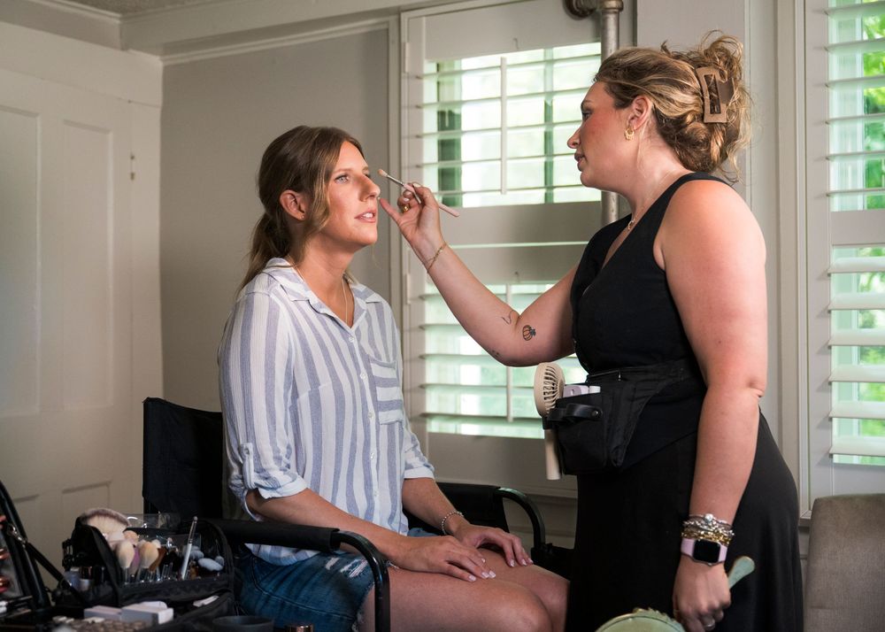 Professional makeup artist applying cosmetics to a client seated in a bright room with window blinds in the background.
