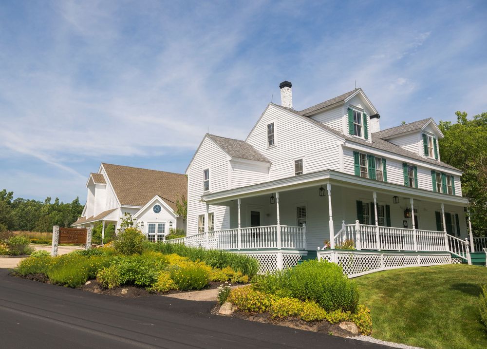 Beautiful white farmhouse style home with wraparound porch and colorful garden landscaping under blue summer sky.