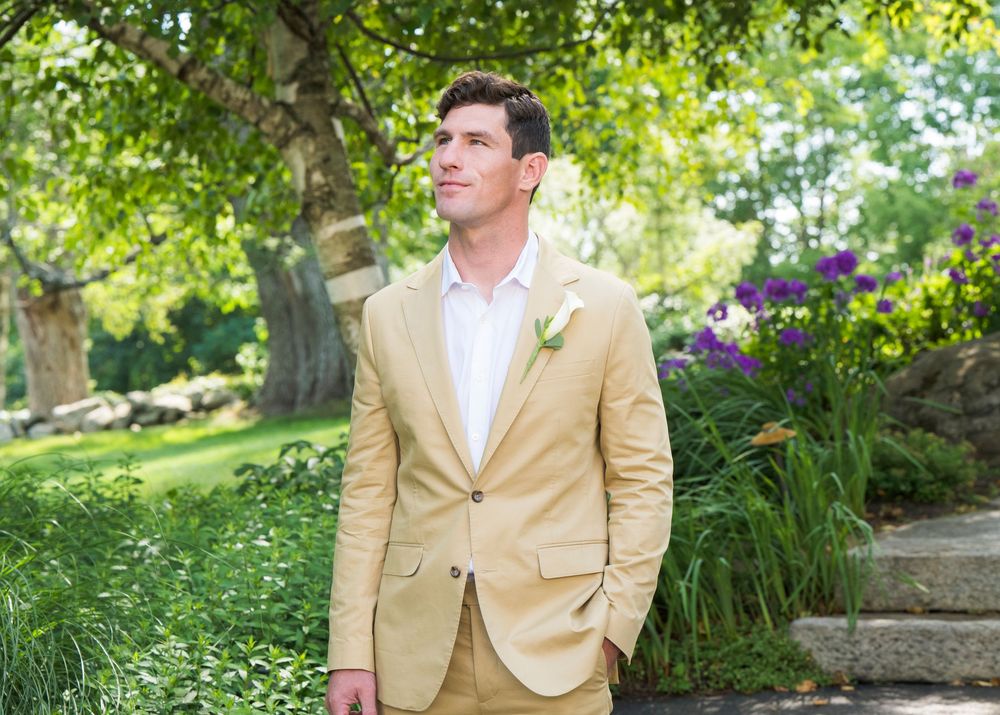 A person in a tan suit jacket and white shirt stands in a lush garden with trees and flowers in the background.