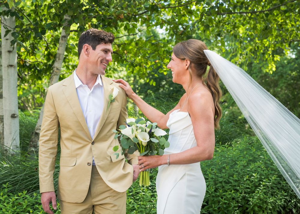 A bride in a white gown and groom in tan suit share a playful moment outdoors under green trees.