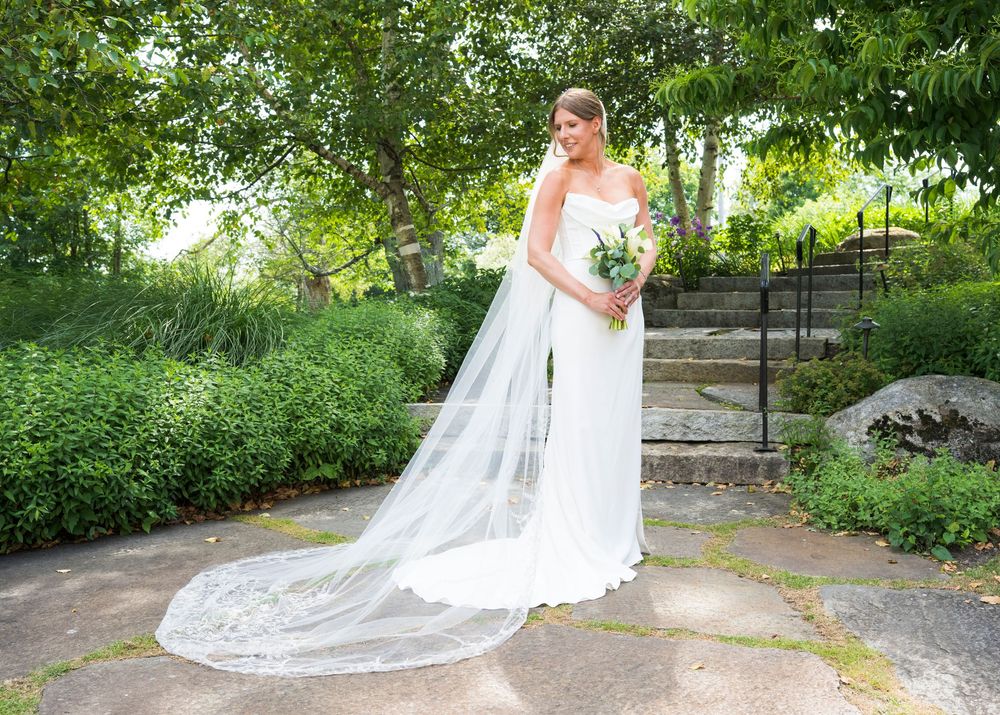 A bride in a white wedding gown with a flowing cathedral veil poses in a lush garden setting on a summer day.