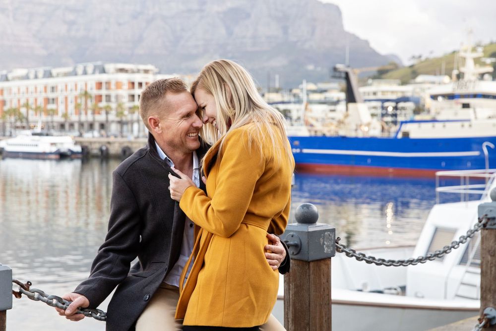 A couple embraces on a dock at a marina with boats and mountains visible in the background.