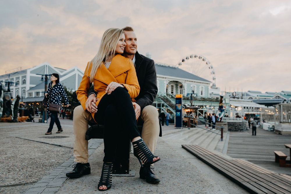 A couple sits on waterfront steps against an urban backdrop with a Ferris wheel at dusk.