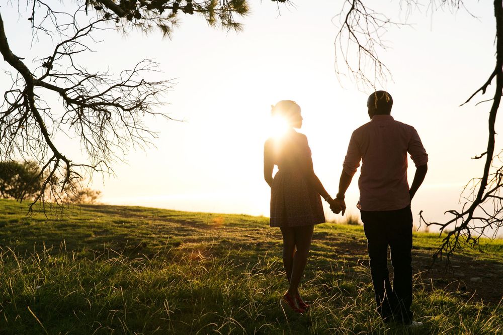 Two silhouetted figures holding hands while walking together against a glowing sunset on a grassy hilltop.
