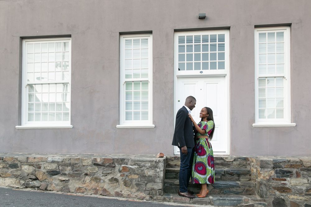 Couple poses against pink stucco wall with stone foundation and white windows, creating a romantic urban backdrop.