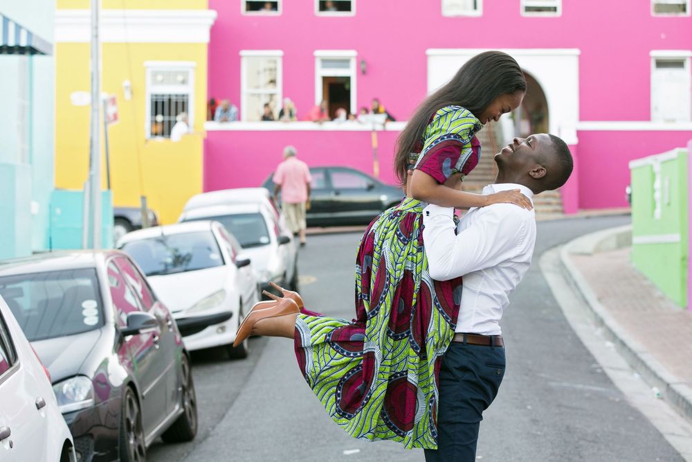 Two people embracing in a street with colorful pastel buildings and parked cars in Cape Town's Bo Kaap neighborhood.