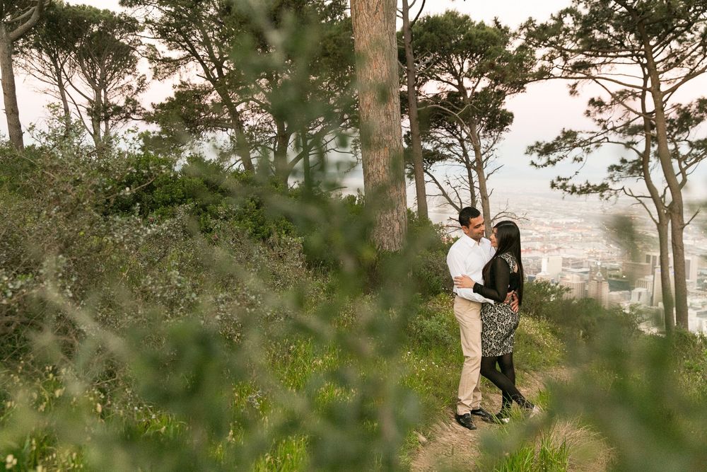 A couple shares a romantic embrace on a scenic hillside path surrounded by tall pine trees at sunset.