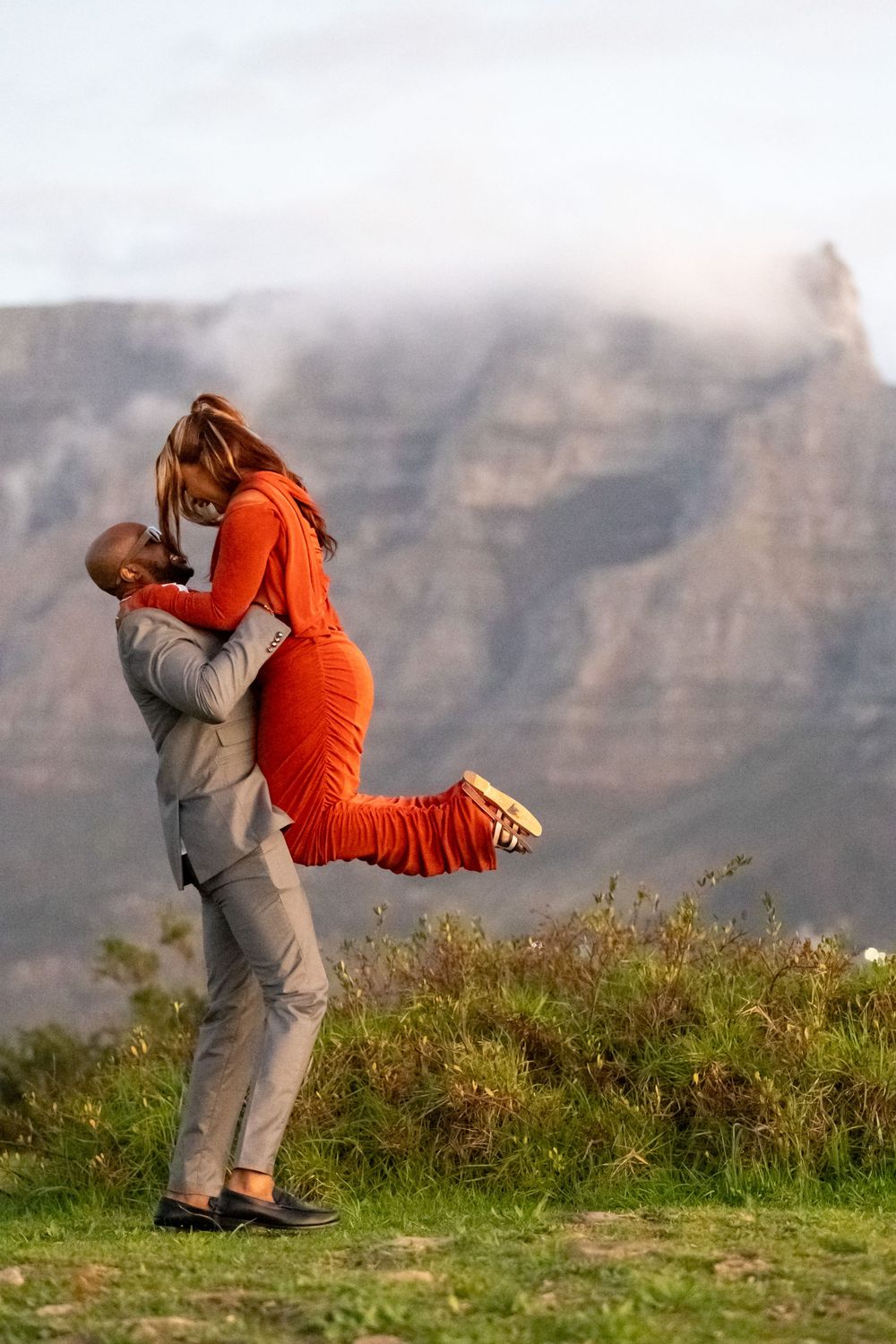 A couple in grey and orange clothing embrace and dance gracefully against a backdrop of misty mountains at sunset.